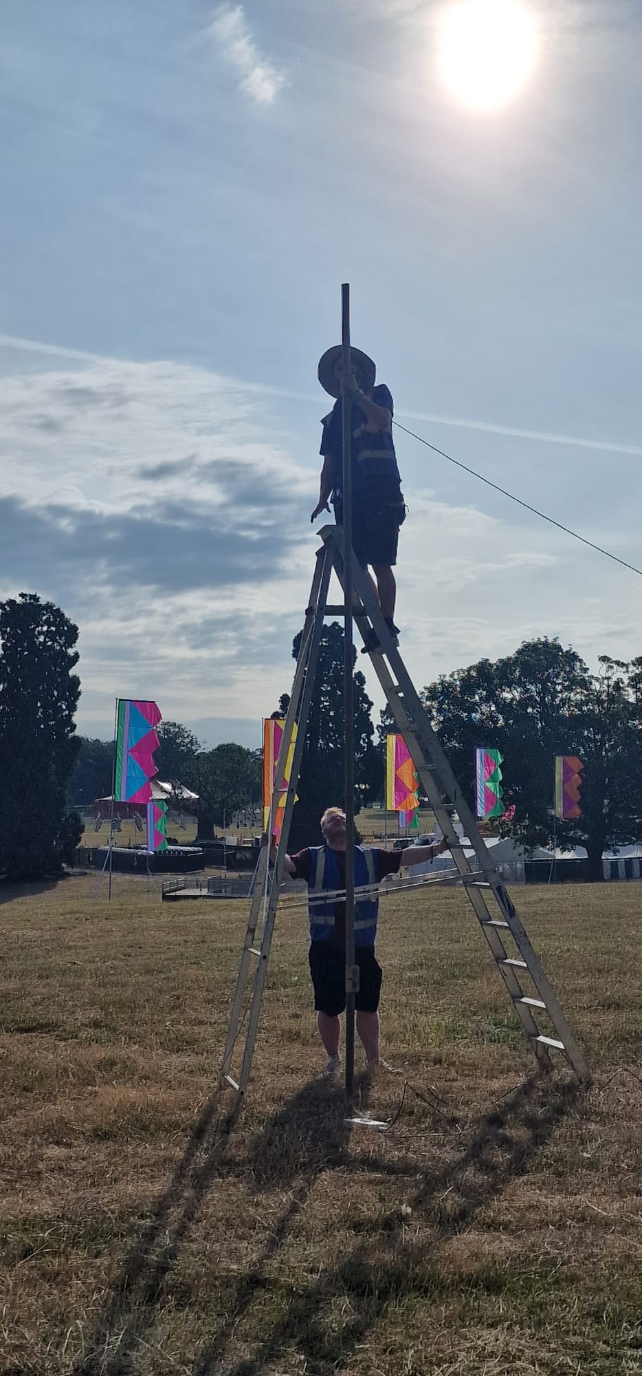 Two HTG engineers working together to raise a mast at Deershed Festival 2026 with festival flags and stage in the background